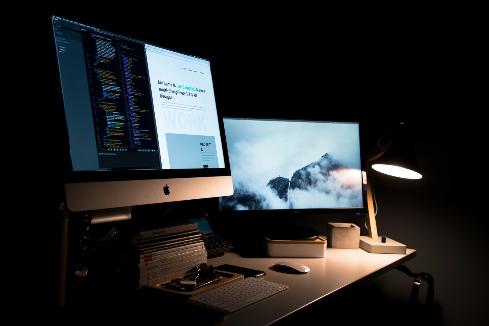 A desk at night with two monitors, one showing code and the other a website design